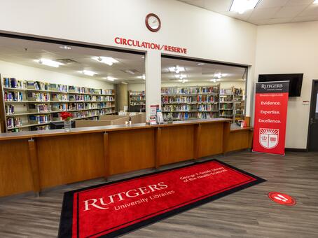 Image of the circulation and reserve desk at the Smith library, with no staff in the image, but stacks of books behind the counter. There is a large red mat in front of the counter that reads "Rutgers University Libraries" as well as lettering above the counter stating Circulation/Reserve