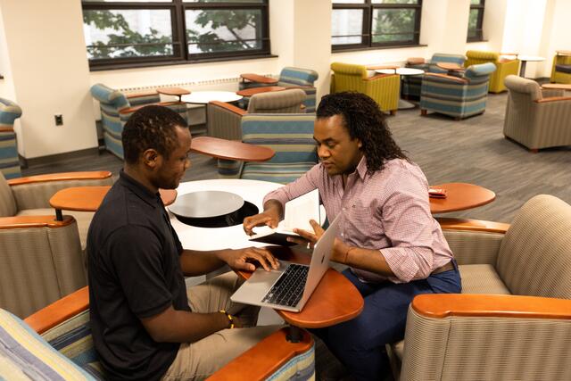 A Robeson faculty member aids a student in the collaborative study space using a book and a laptop