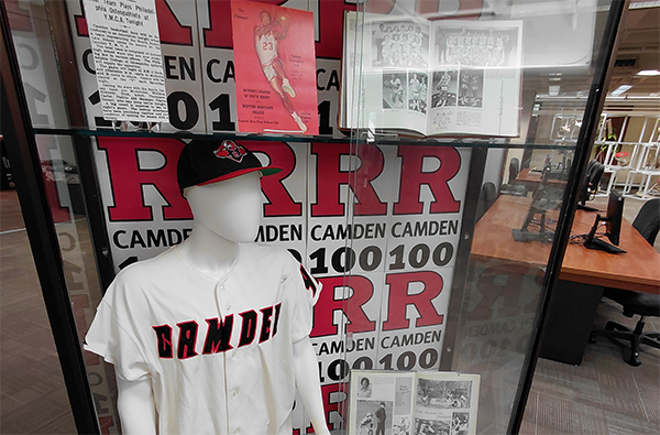 Image of an exhibit case featuring Rutgers-Camden sports memorabilia, including a baseball jersey, year books, and sporting events programs 
