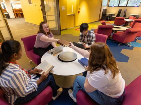 Four students in a study space, two are passing a book across a white circular table
