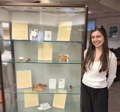 Image of student artist Sophia Westfall standing next to a display case filled with her work.