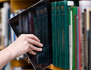 Image of a hand pulling an electronic tablet off a bookshelf, the bookshelf is filled with physical books