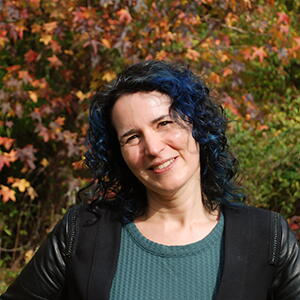 Photo of Caryn Radick in front of foliage, she has blue hair and is wearing a black jacket with a light green top