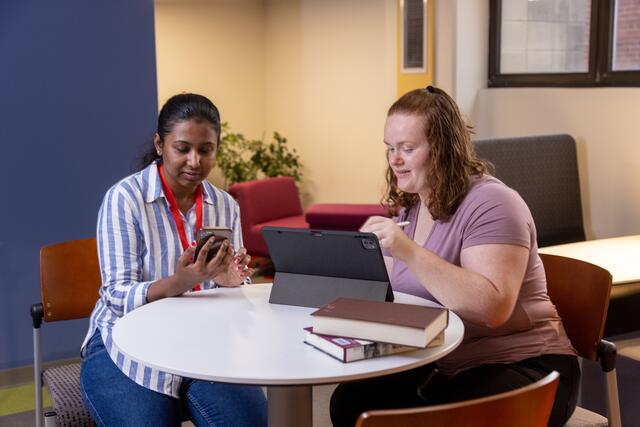 Photo of students working together at a round, white table. They are working with a laptop and a smartphone, with a short stack of books with them.