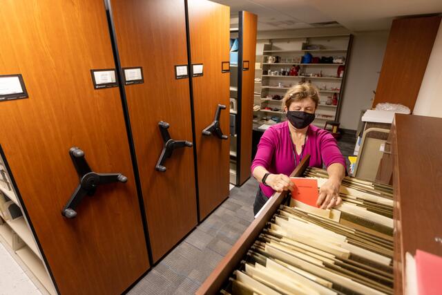 Image of an archivist taking a document from the collections drawers. She is wearing a black mask and a pink sweater. The moveable shelves can be seen behind her