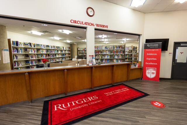 Image of the circulation and reserve desk at the Smith library, with no staff in the image, but stacks of books behind the counter. There is a large red mat in front of the counter that reads "Rutgers University Libraries" as well as lettering above the counter stating Circulation/Reserve