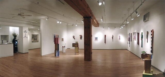 Large art gallery room with wall art and floor sculptures. A thick wood beam spans the length of the main gallery and connects to a thick wood vertical column. The gallery contains many lights which are on.
