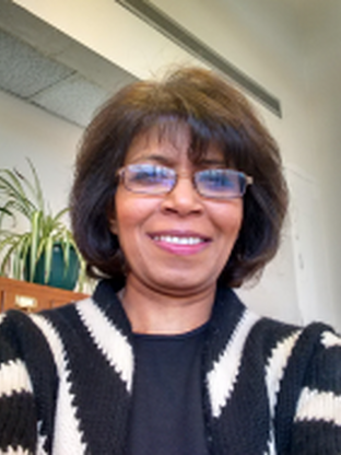 A photo of Kalaivani, she is wearing glasses and a black shirt with a black and white striped cardigan over the shirt. In the background is a shelf with a plant on it 