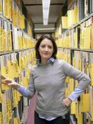 Photo of Adriana in between two bookshelves with yellow files. She is wearing a beige sweater and dark pants