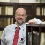 Photo of Robert wearing a white dress shirt and a red tie, leaning against a book shelf with more book shelves in the background
