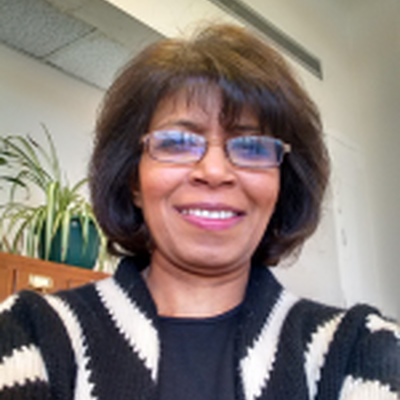 A photo of Kalaivani, she is wearing glasses and a black shirt with a black and white striped cardigan over the shirt. In the background is a shelf with a plant on it 