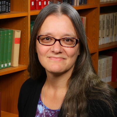 Photo of Stephanie Bartz, she is wearing glasses and a black sweater. She is standing in front of bookshelves