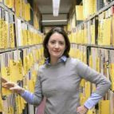 Photo of Adriana in between two bookshelves with yellow files. She is wearing a beige sweater and dark pants