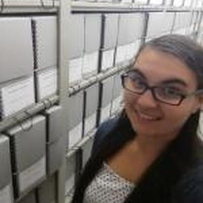 Photo of Tara in front of bookshelves, she is wearing glasses and a white tank top with a black cardigan over the tank top.