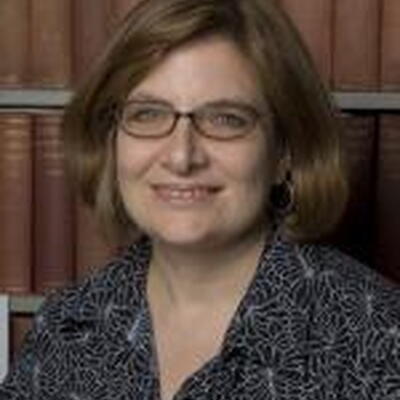 Photo of Fernanda, wearing a dark patterned shirt and glasses standing in front of book shelves