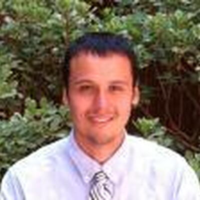 Photo of Jonathan, wearing a white dress shirt and a tie in front of hedges