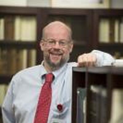 Photo of Robert wearing a white dress shirt and a red tie, leaning against a book shelf with more book shelves in the background