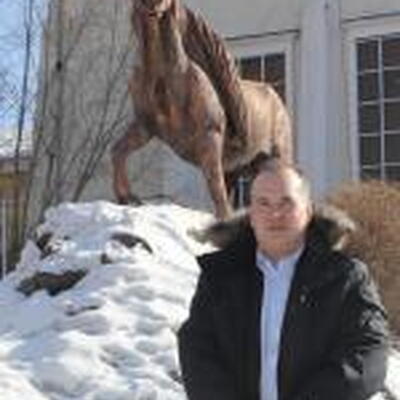 Photo of Ryan standing in front of a horse statue with snow at the horse's feet. Ryan is wearing a black jacket over a light colored shirt.