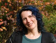 Photo of Caryn Radick in front of foliage, she has blue hair and is wearing a black jacket with a light green top
