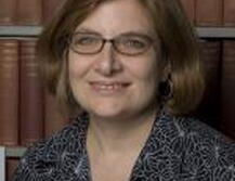 Photo of Fernanda, wearing a dark patterned shirt and glasses standing in front of book shelves