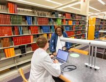 A student studying at one of the open desks. Behind him are the stacks of books