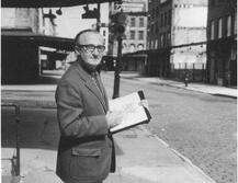 Image of John de Pol, standing on a street corner while holding a book and pen. The photo is black and white